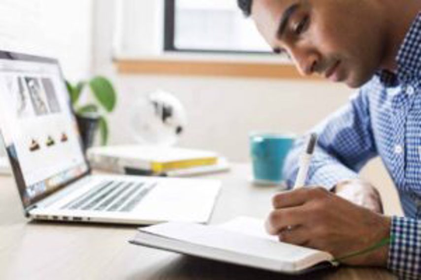 A man writes in a notebook while sitting at his computer