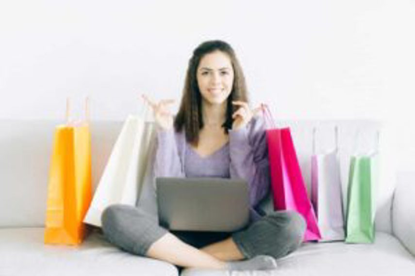 A woman sits on her couch with her computer on her lap and shopping bags around her